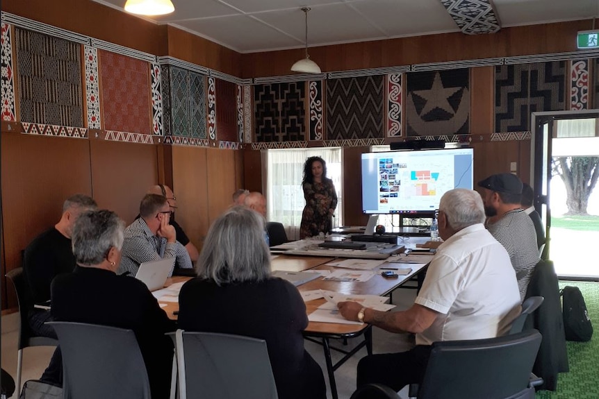 Woman stands presenting with screens behind her in marae with people sitting around the table.