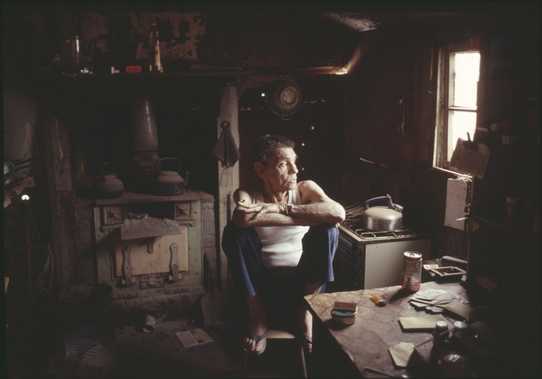 A man in a singlet sits in his home and looks out the window. 