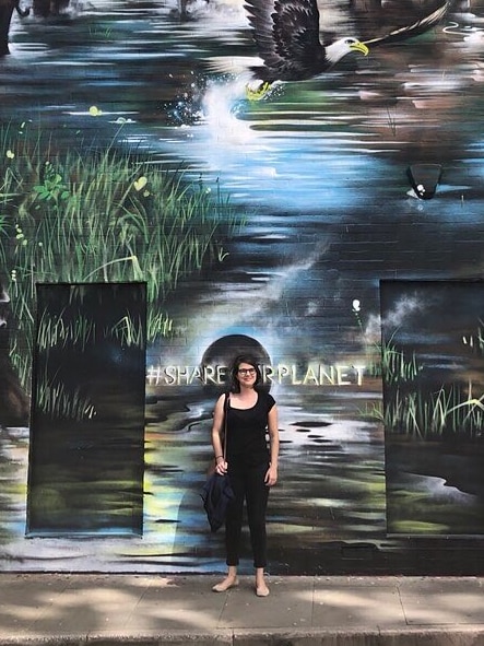 A young woman poses smiling on the sidewalk next to a mural.