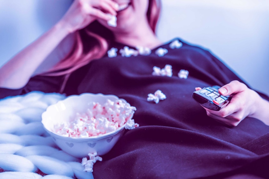 A woman lying back eating popcorn with a TV remote in her hand