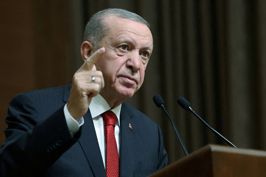 An older Turkish man in a suit speaks on a podium, pointing to the air