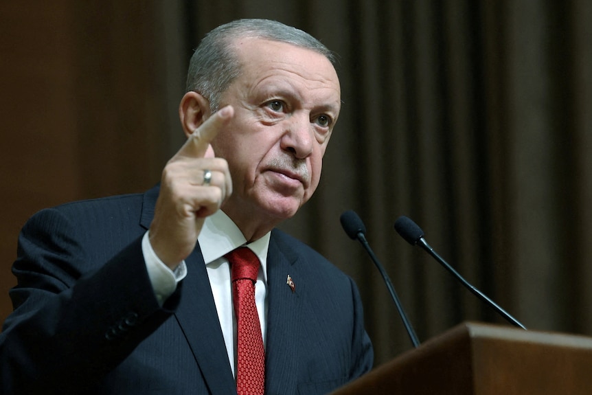 An older Turkish man in a suit speaks on a podium, pointing to the air.
