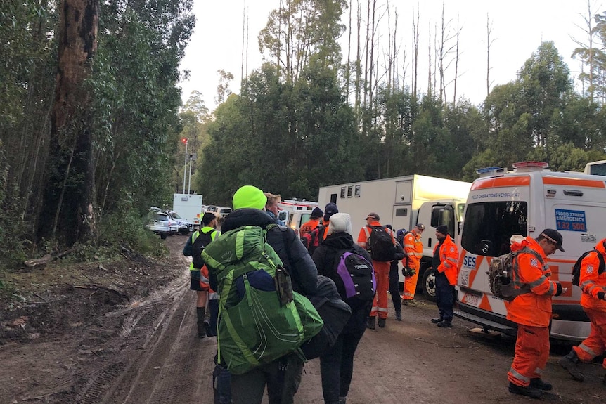 People wearing hiking gear and backpacks stand around SES cars and trucks in the carpark of a state forest.