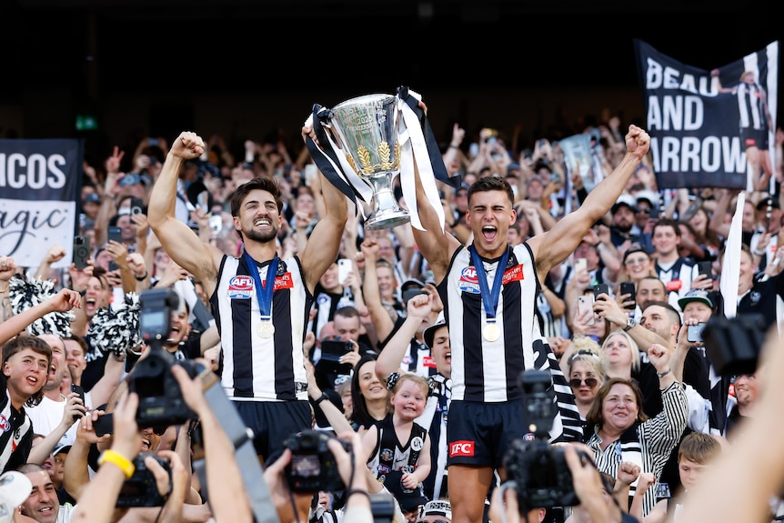 AFL players, and brothers, holding the premiership cup while celebrating with thousands of fans