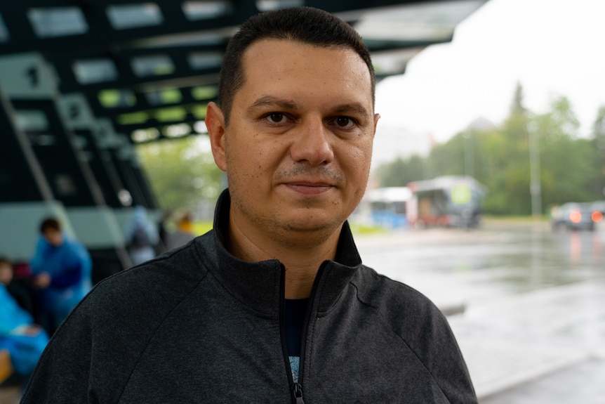 A man with dark hair in a black sweater poses for a photo at a bus station.