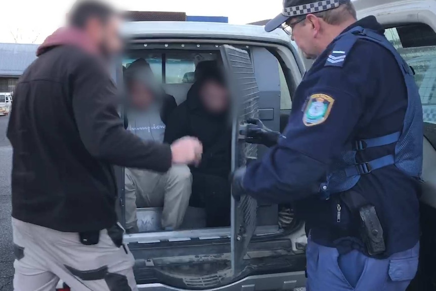 Two men, whose faces are blurred, are placed in the back of a police vehicle.