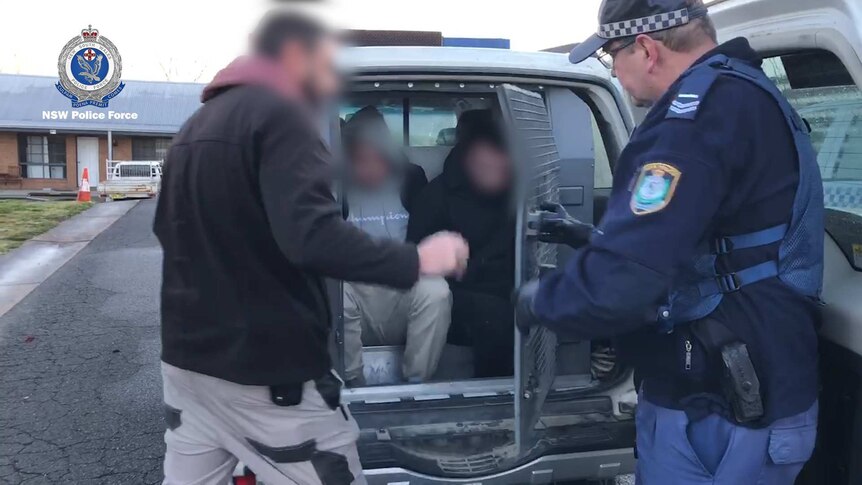 Two men, whose faces are blurred, are placed in the back of a police vehicle.