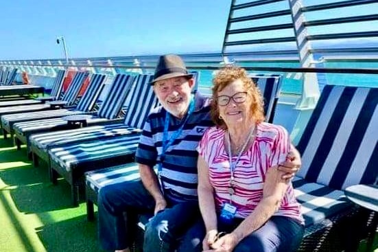 Jane and John Newson pose sitting down, smiling, on a cruise ship, with ocean in the background.
