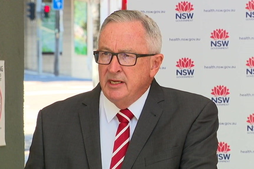 A man with a red and white striped tie stands in front of a background with NSW Government logos