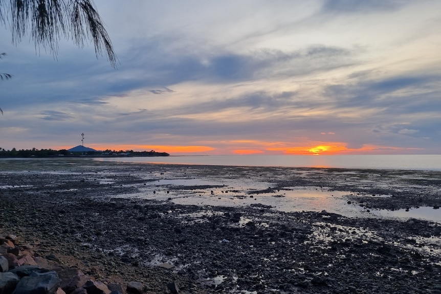 An orange sunset in a cloudy sky over the rocky shoreline of Saibai 