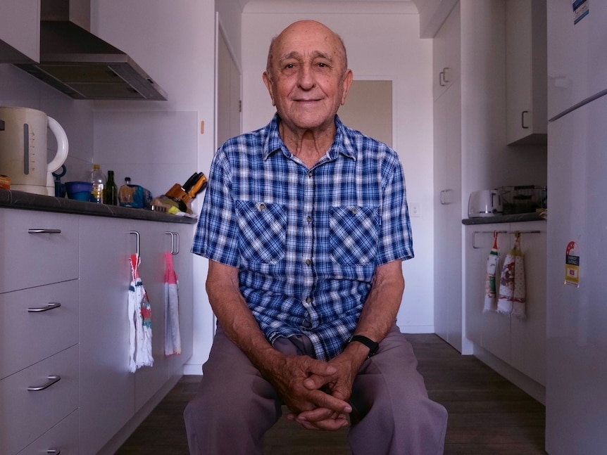 An elderly man sits in his kitchen on a chair in a blue check shirt