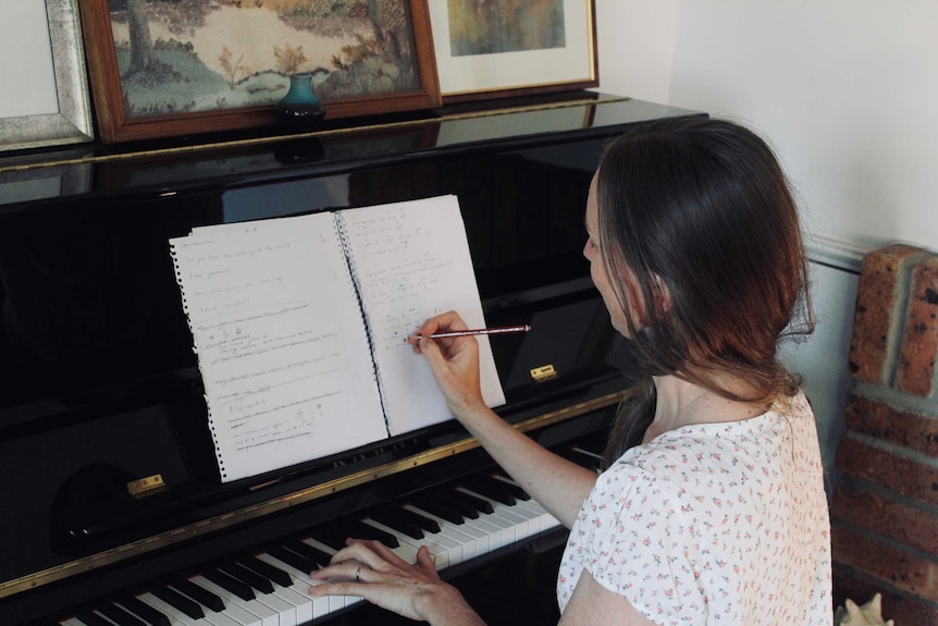 A woman with dark hair sits at a piano writing on a sheet of music.