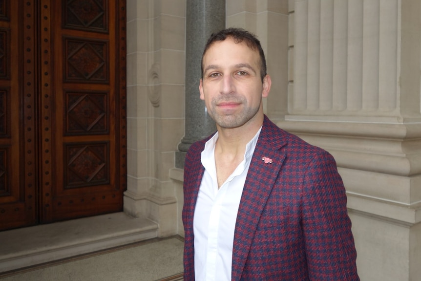 Man in white shirt and checked blazer looks seriously at camera in a courtyard