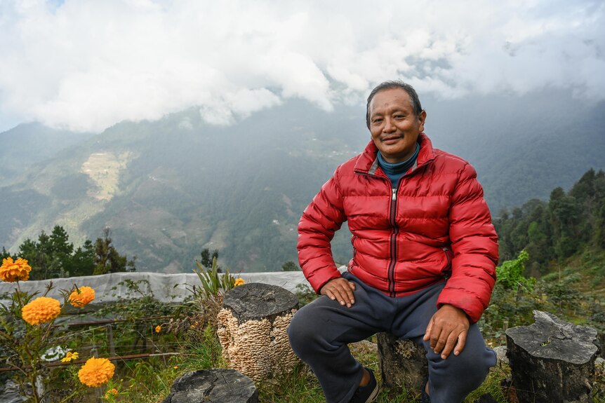A man in a red puffer jacket sits on a tree stump with a mountain valley behind him