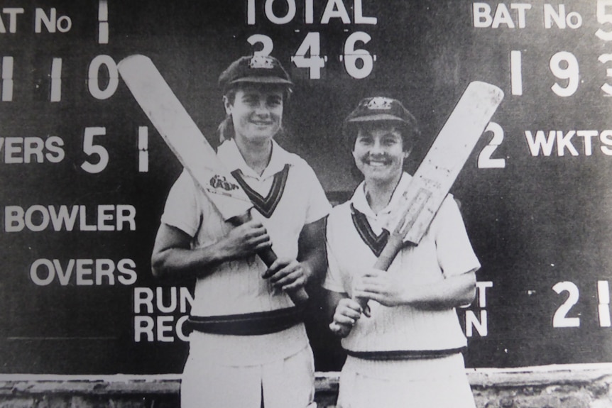 Lindsay Reeler (left) and Denise Annetts (right) in front of a scoreboard holding cricket bats.