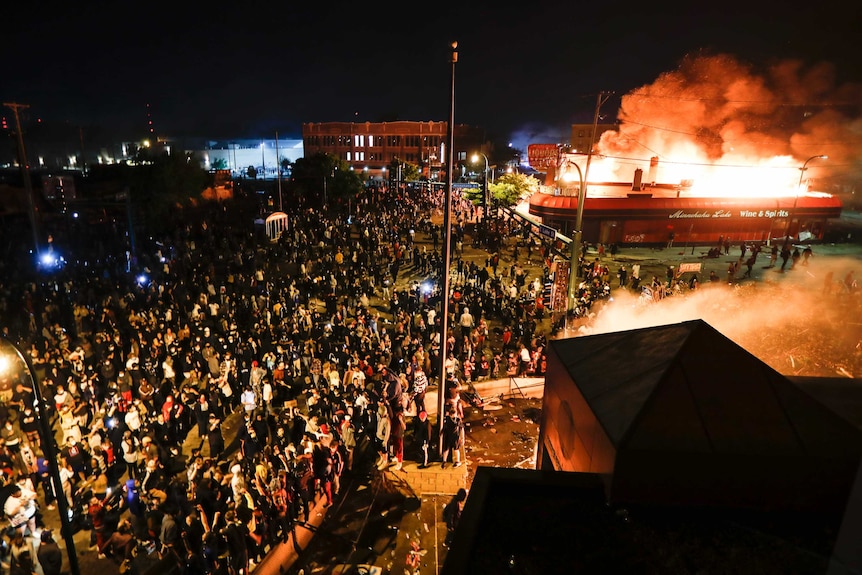 Hundreds of people stand and watch as a police station burns. The image is shot from above, and night has fallen.