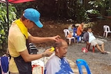 A man wearing yellow trims another man's hair outside as others watch on.