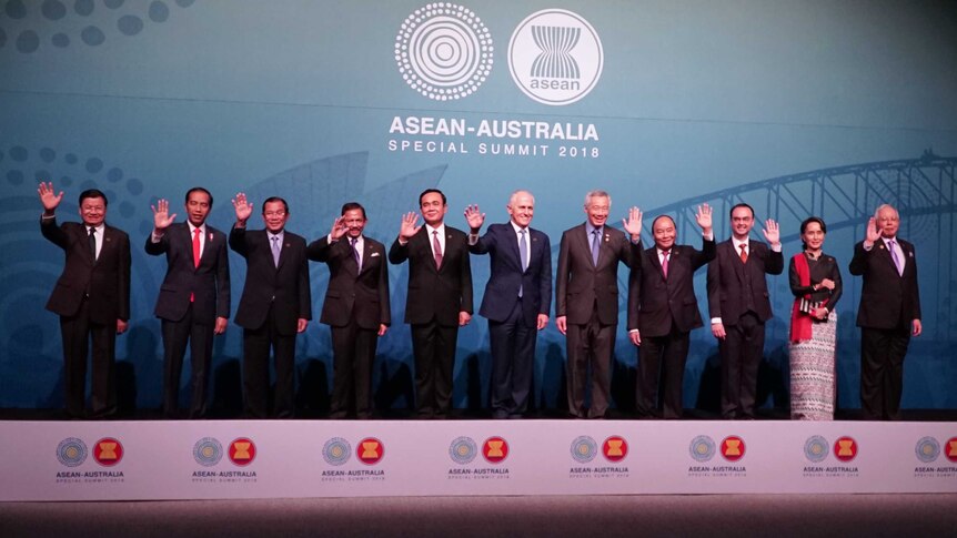 Politicians stand in a line waving in front of a blue background.