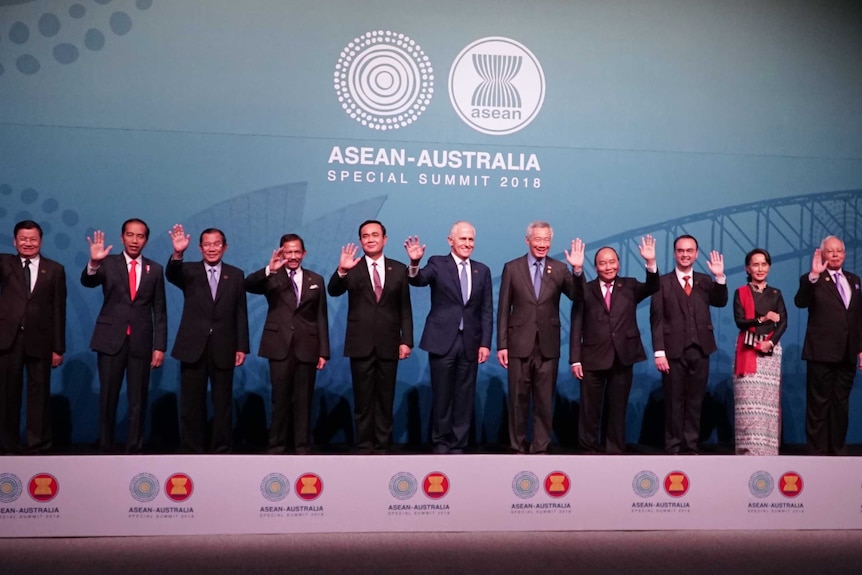 Politicians stand in a line waving in front of a blue background.