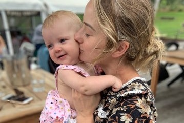 Women holds baby wearing pink jumpsuit in her arms and kisses her on the cheek.