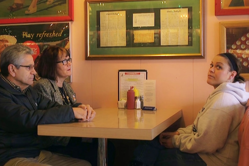 A thirty-something woman sits opposite her parents in a diner booth, looking fed up as they look at her. 