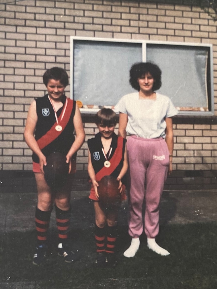Two boys stand next to each other in Aussie rules football gear and medals around their neck, with their mum next to them.