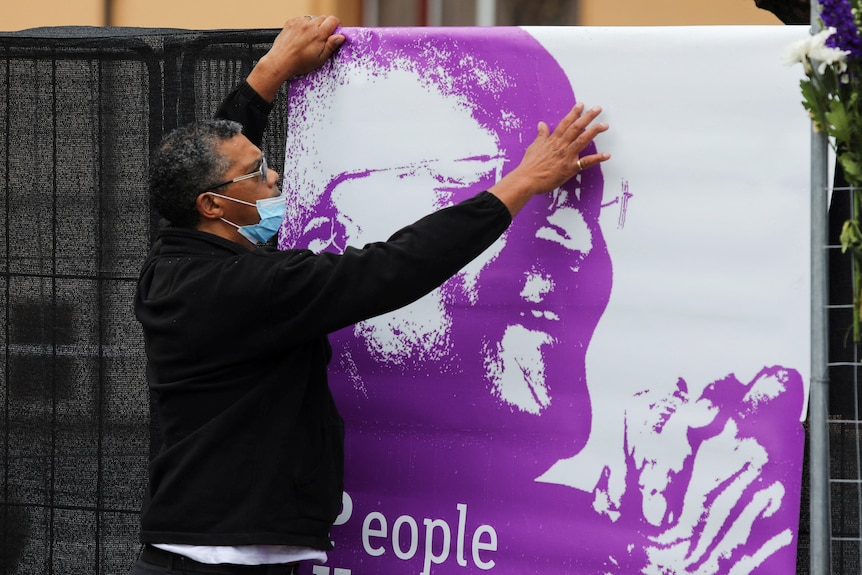 A middle-aged man in a face mask hangs a white and purple poster of Desmond Tutu at his funeral