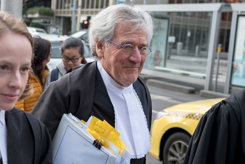 A man with grey hair and wire-rimmed glasses walks on the street wearing black and white legal robes