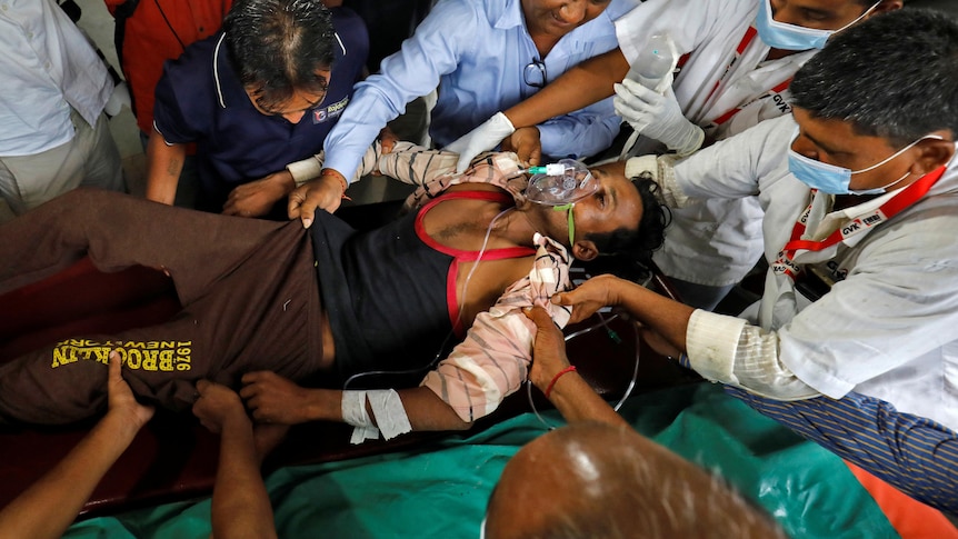 A semi-conscious young Indian man wears oxygen mask and is surrounded by people as he is carried