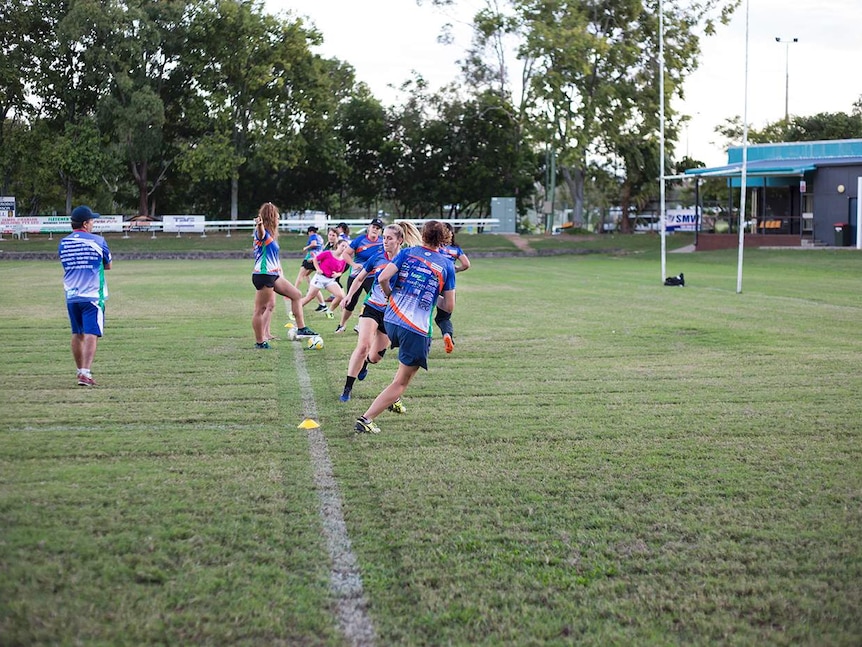 Row of girls runs towards the line on a field, and directed by another player who has her arm outstretched, foot resting on ball