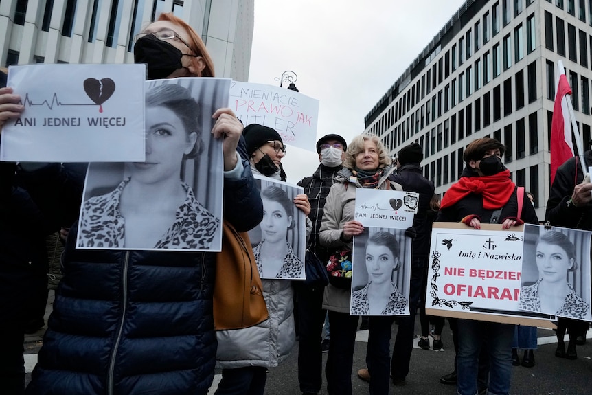 Protesters standing among tall buildings hold black and white photos of a woman who died of complications from her pregnancy.