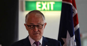 Malcolm Turnbull, wearing glasses, looks down towards a lectern. Behind him is a green EXIT sign and an Australian flag.