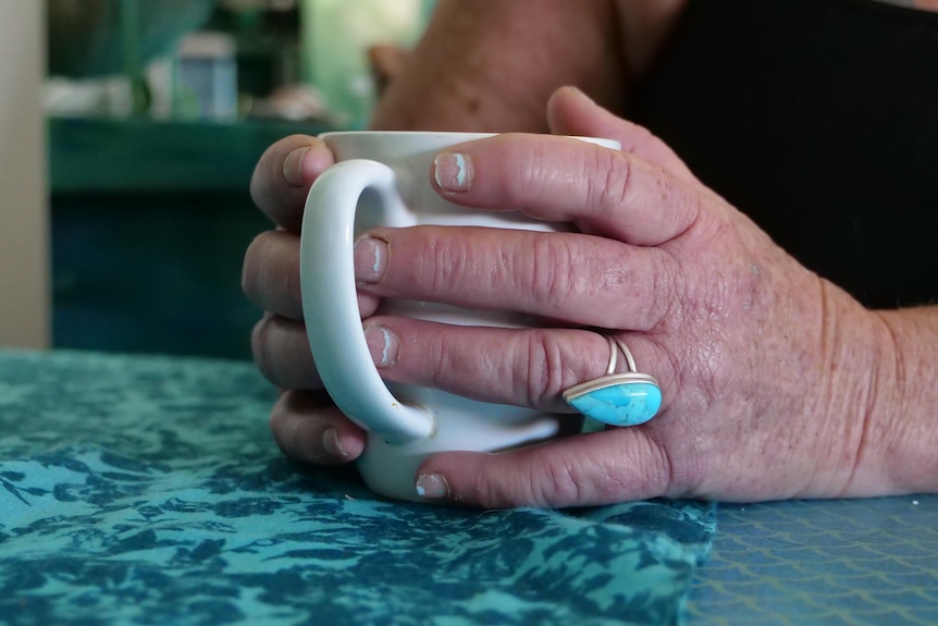 Two hands hold a coffee mug, one of her fingers has a big blue ring on, she's sitting at a blue-coloured table.