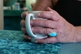 Two hands hold a coffee mug, one of her fingers has a big blue ring on, she's sitting at a blue-coloured table.