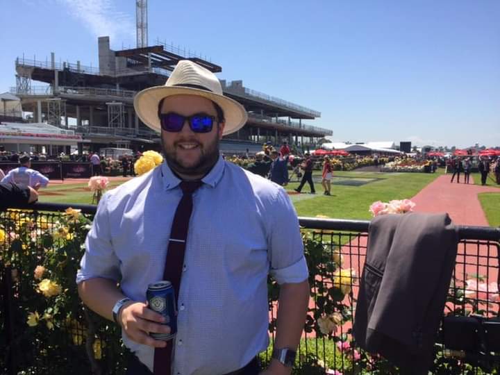 A man in a hat and glasses stands in the sunshine at a racetrack