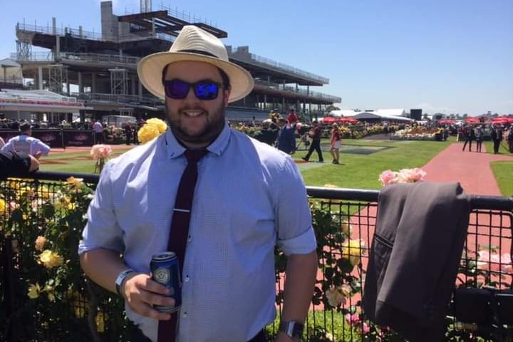 A man in a hat and glasses stands in the sunshine at a racetrack