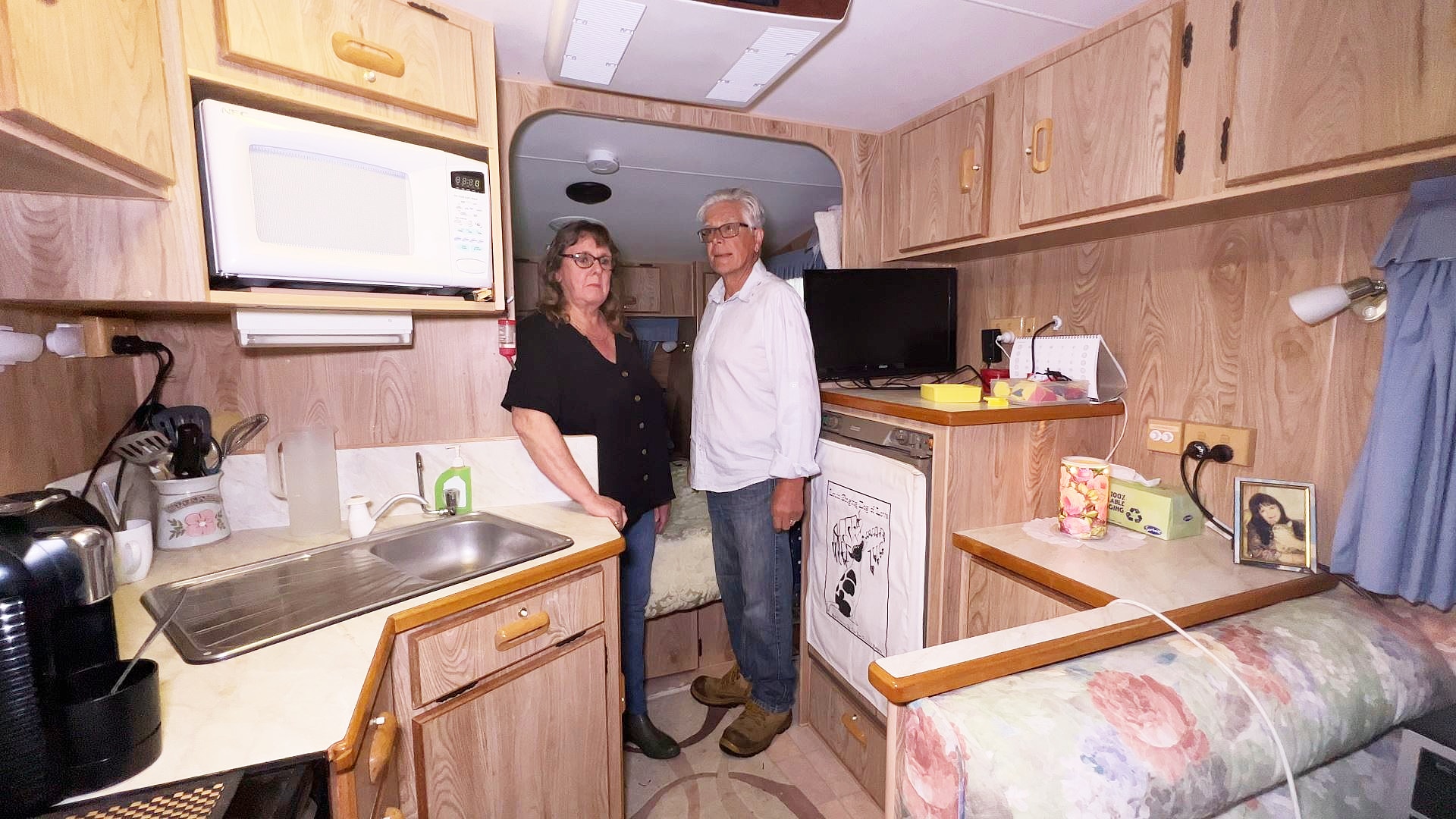 A couple stand in a caravan surrounded by a workspace and fridge.