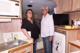 A couple stand in a caravan surrounded by a workspace and fridge.