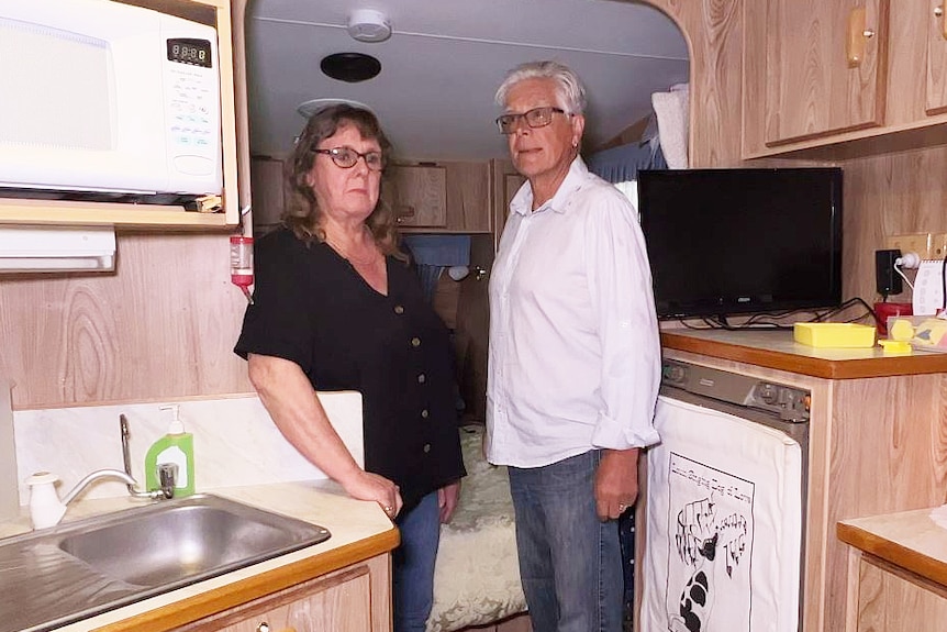 A couple stand in a caravan surrounded by a workspace and fridge.