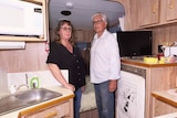 A couple stand in a caravan surrounded by a workspace and fridge.