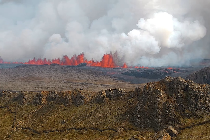 Bright orange lava spews into the sky amid a green, mountainous landscape