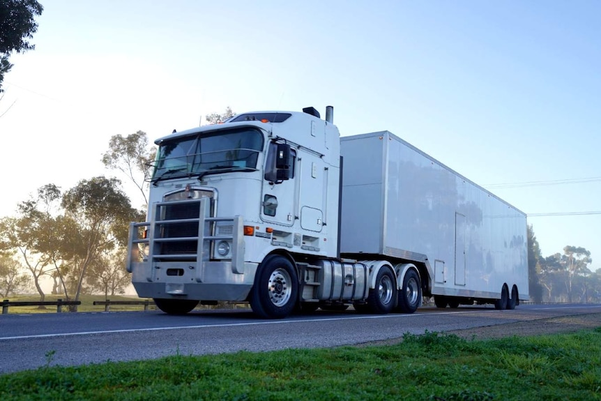 A white truck drives a long a highway at a golden sunrise, passing large gum trees.