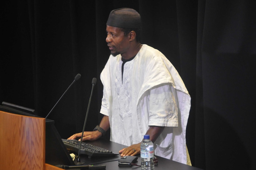 Imam Adama Konda in white robes, speaking at a podium.