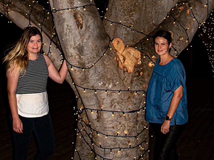 Two women stand next to a tree covered in fairy lights