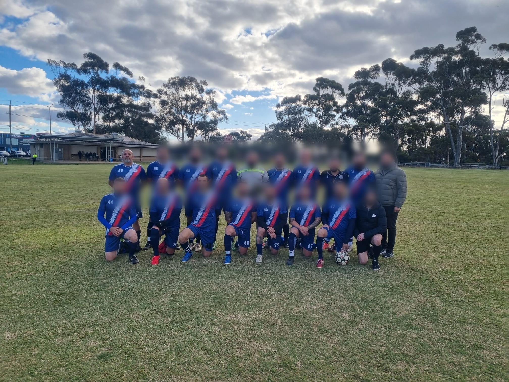 A bald man stands in a group photo on a field with men in blue soccer uniforms wiht a white and red diagonal stripe.
