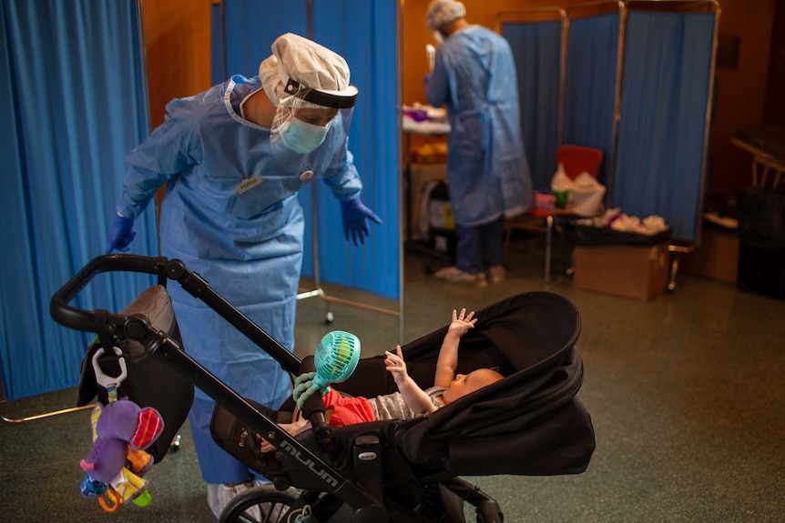 A volunteer dressed in hospital gear and face mask plays with a 4-month old baby on a pram.