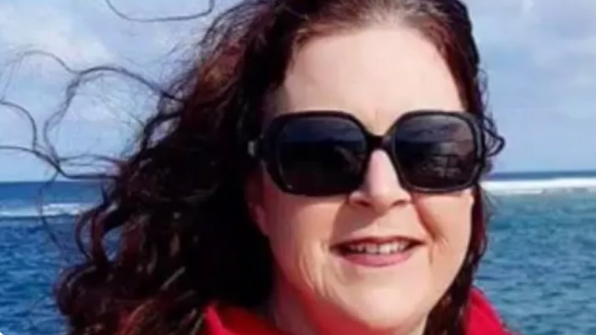 A woman with dark red hair and sunglasses smiles at the camera. She is at a beach.