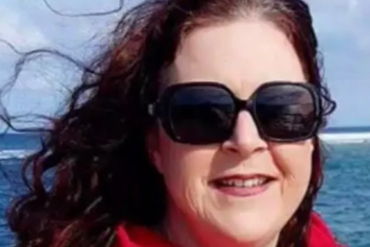 A woman with dark red hair and sunglasses smiles at the camera. She is at a beach.