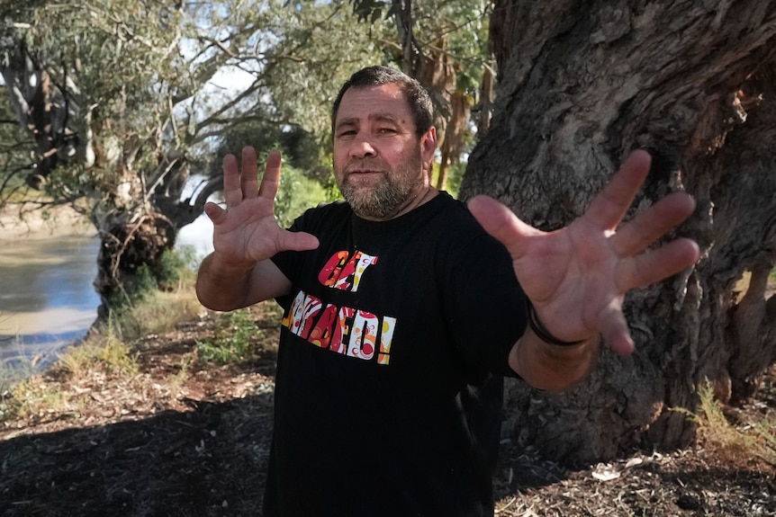 A man with a "get jabbed" T-shirt with his hands out, on a riverbank.