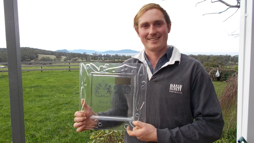 A smiling young man holding a glass award for his dairy at Bream Creek producing the best milk in the Tasmanian Fine food awards
